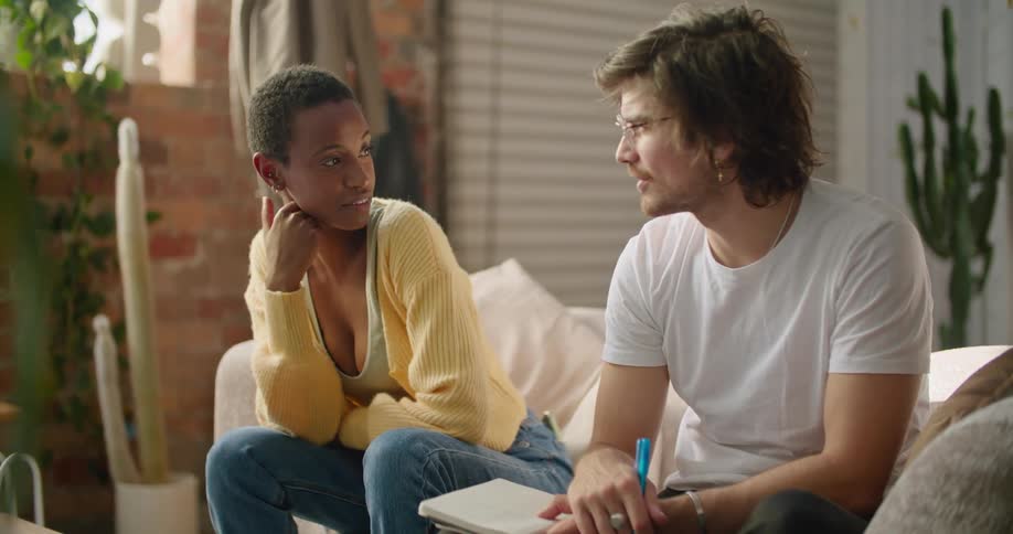 A man and a woman sitting in a room talking with a wall and window in the background
