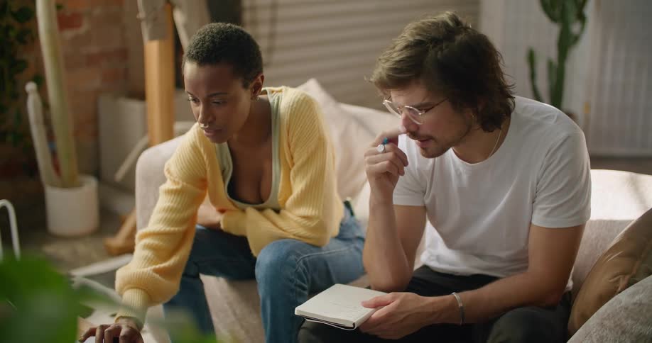 A man takes notes while watching his friend scroll through a laptop