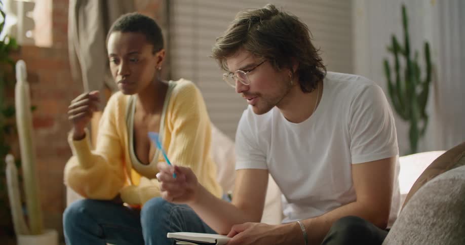 A man and a woman sitting in a room looking at a laptop with a wall and window in the background