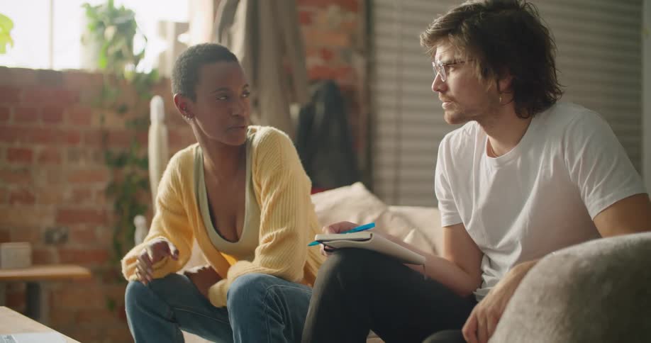 A man and a woman sitting in a room while the woman uses a laptop with a wall and window in the background