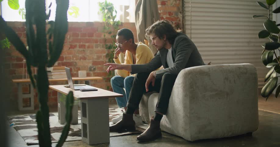A man and a woman sitting in a room looking at a laptop with a wall and window in the background