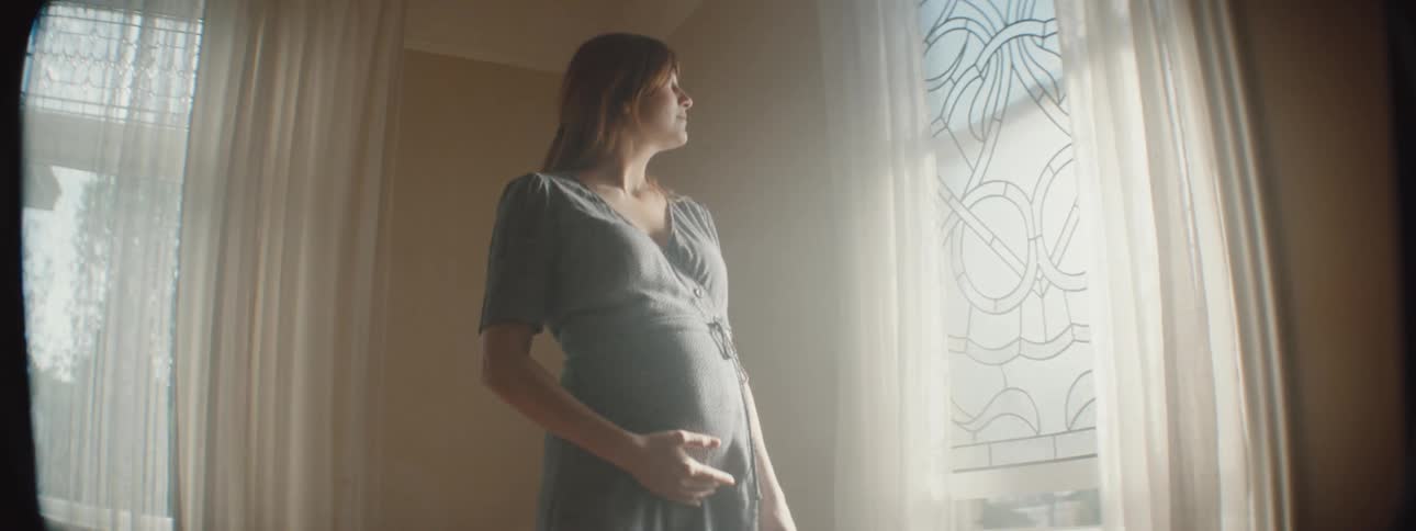 A pregnant woman holds her stomach as she looks out a window and walks around a room in her home