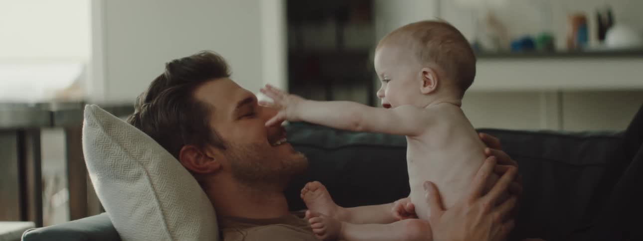 A father reclines on a couch while he lets his infant child play and touch his face