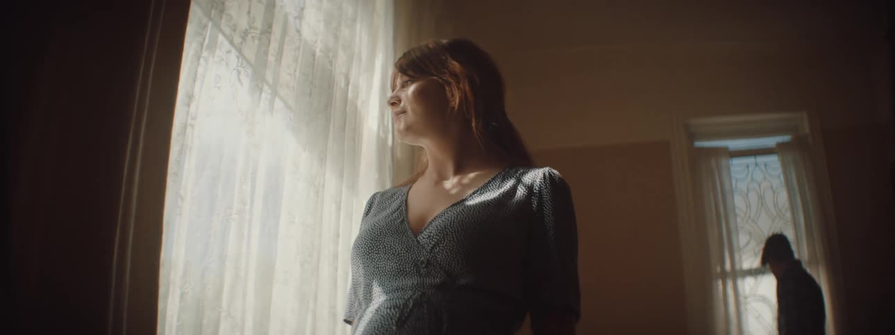 A woman slowly walks through a darkened room and pauses to look at the curtain draped in front of a large window