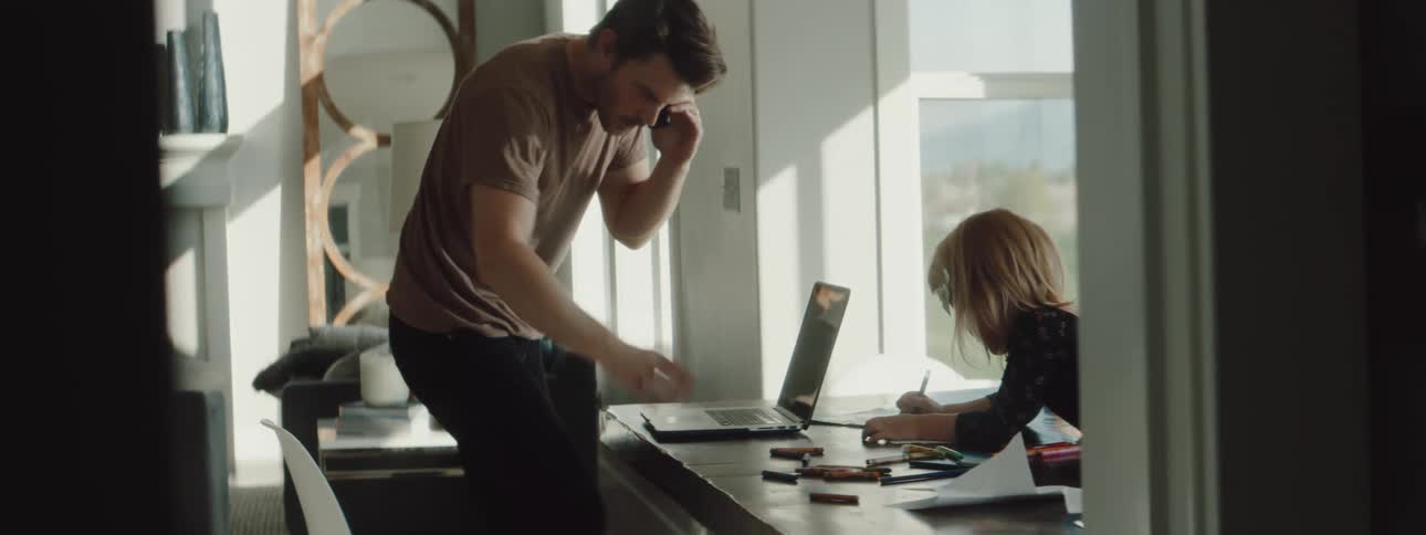 A man is sitting at a table with a laptop and a young girl coloring then gets up from the table talking on a cell phone