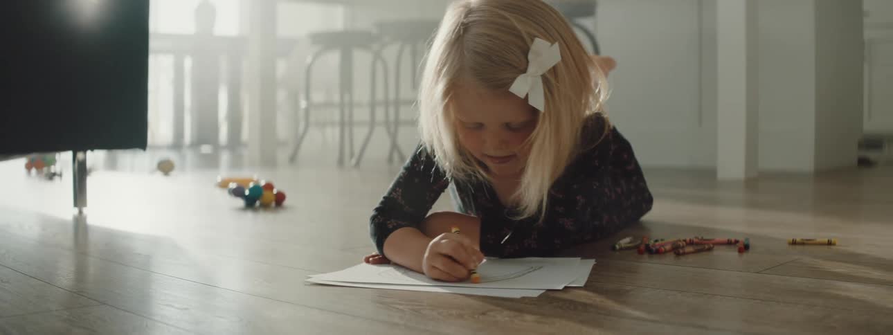 Blonde girl lies on floor coloring as sun shines through glass past furniture