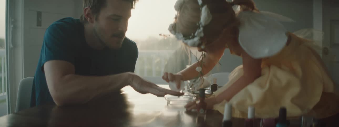 Girl paints her father fingernails while sitting over the table at sunset