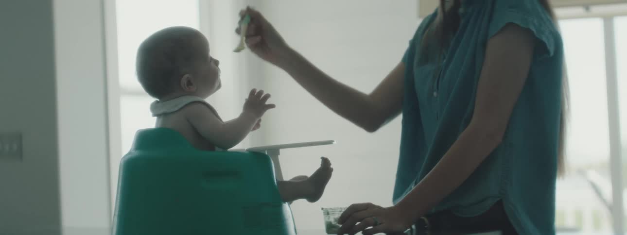 A woman feeding a baby in a high chair with a wall and window in the background