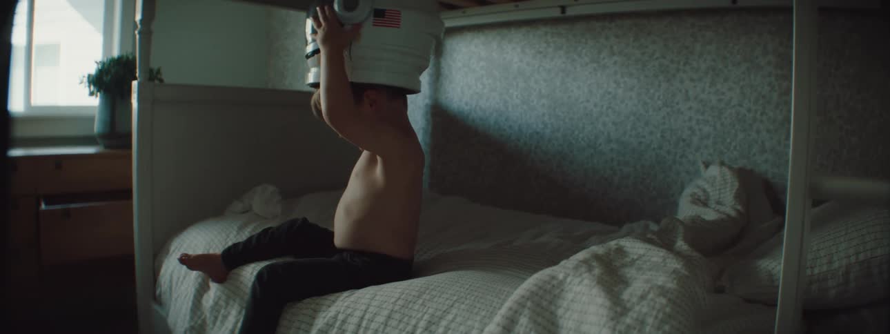 A young boy at home sits on his bed and puts on a space helmet
