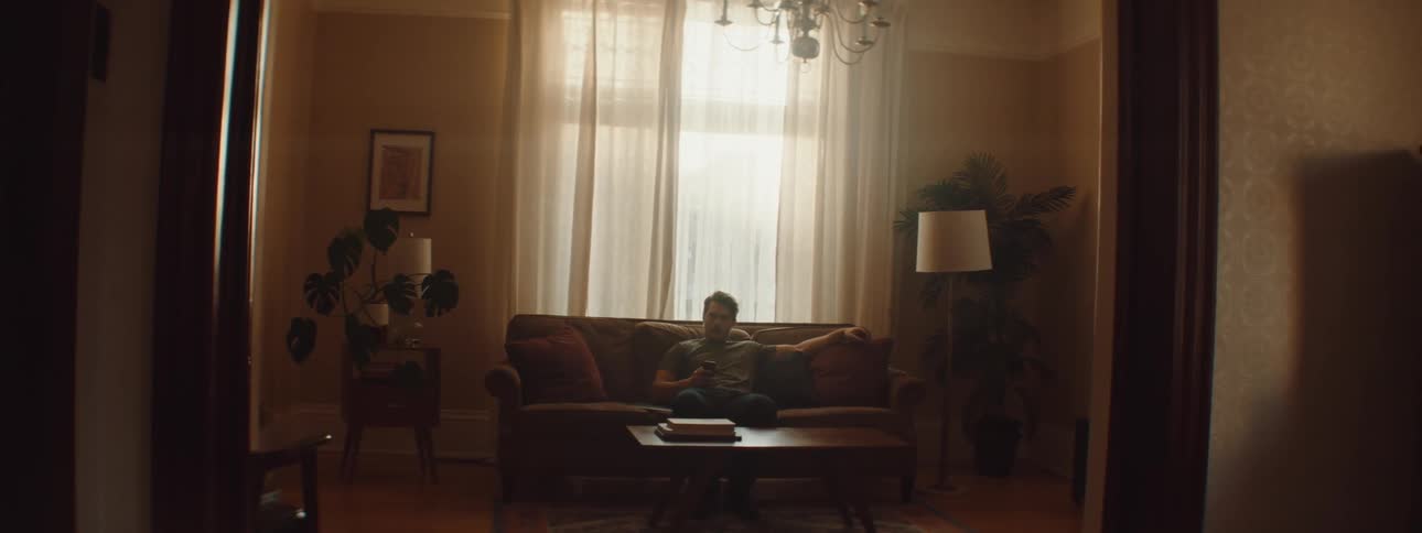 A man sitting on a couch in a living room texting on a cell phone during the day