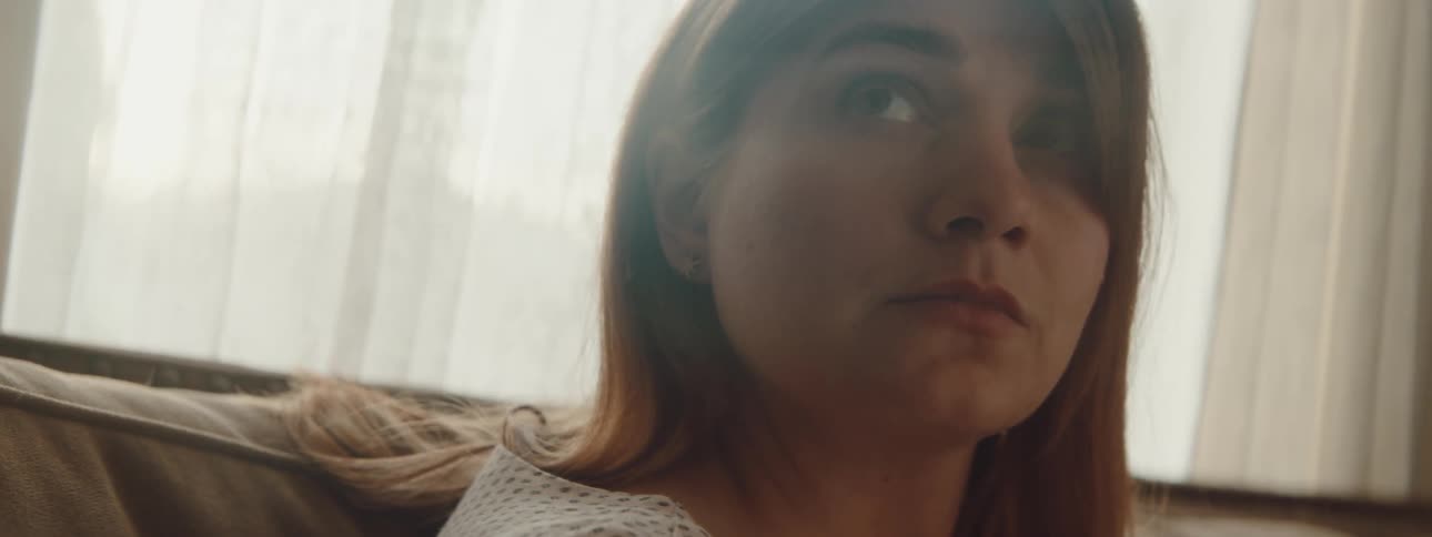 A woman in a room sitting on a couch with a window and blinds in the background