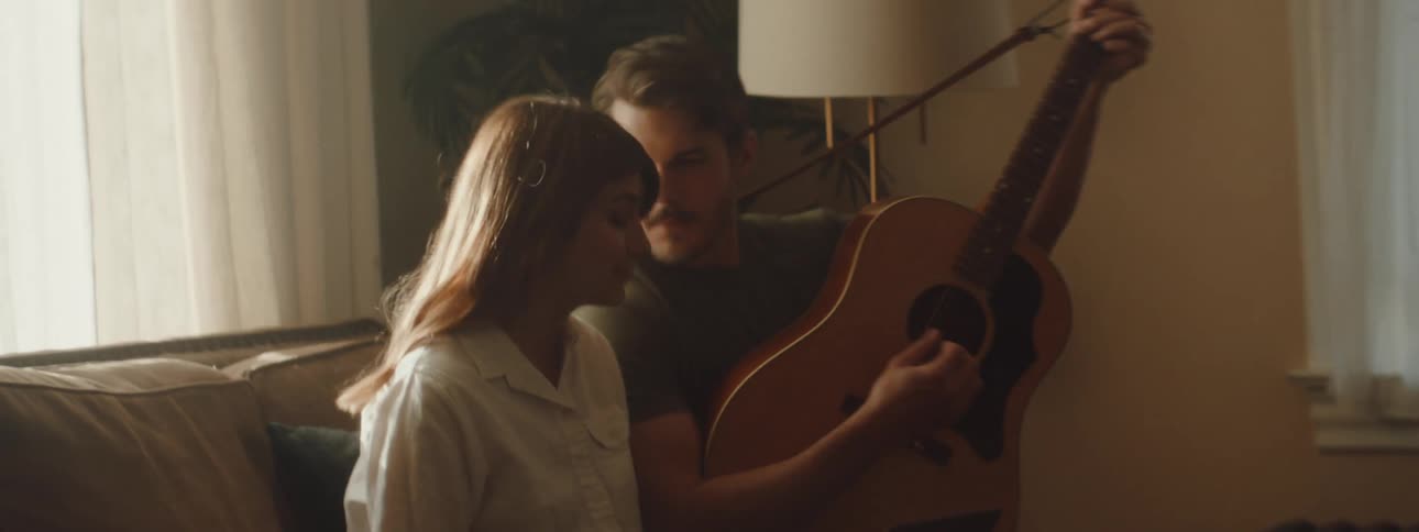 A woman sits on a couch and a man sits down beside her playing the guitar