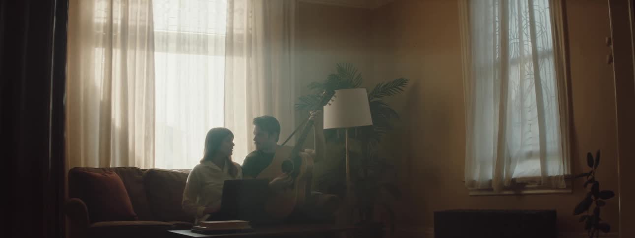 A woman sits on a couch with her laptop and a man runs in the room playing his guitar and singing and sits next to her