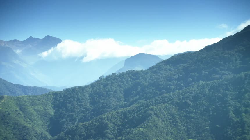 Clouds over a large mountain range covered in green forests under a blue sky