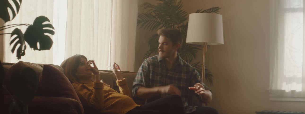 Man with a shirt and a beard sits down on couch next to pregnant woman in turtleneck sweater