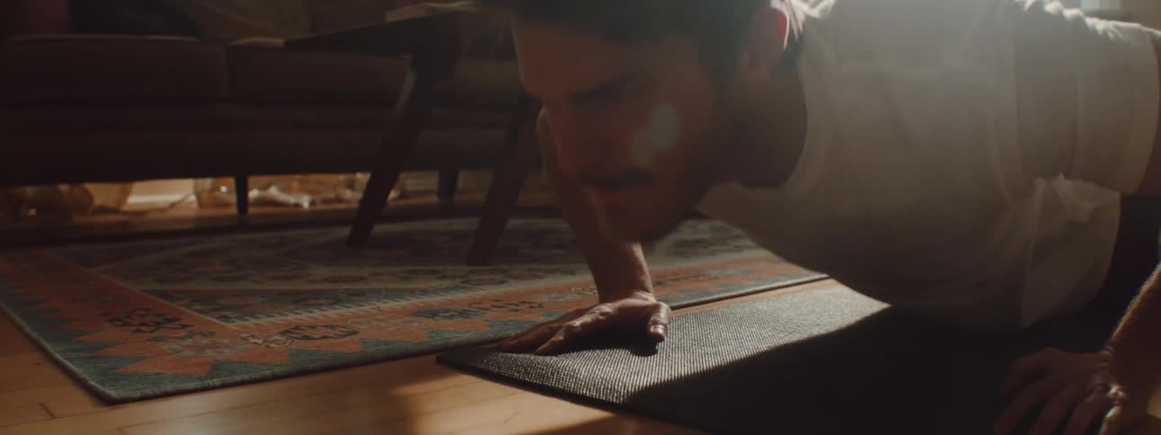 Man with a white undershirt and back shorts does push ups in the living room at home