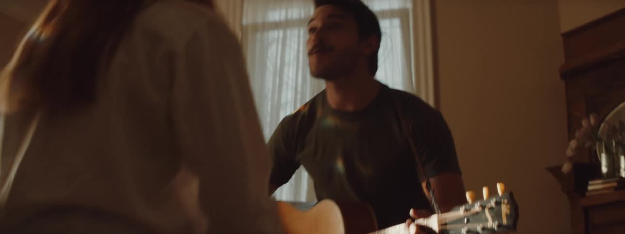 A man plays guitar and a woman jumps around dancing in the living room with him