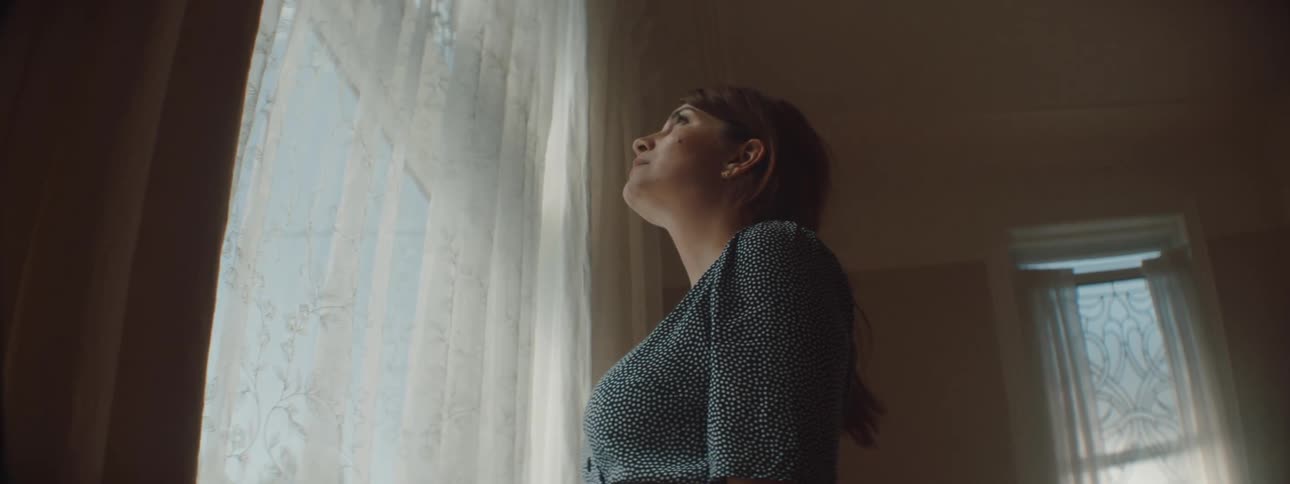 A woman standing before a large window looks up at the curtains hanging in front of it