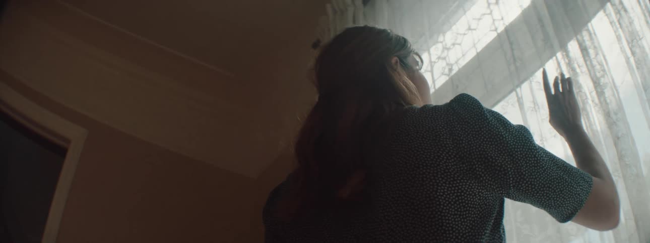 A woman at home parts her drapes and looks out the window