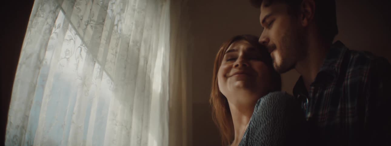 Man with a beard and a shirt approaches woman touching the window curtains and cuddles her in the living room at home