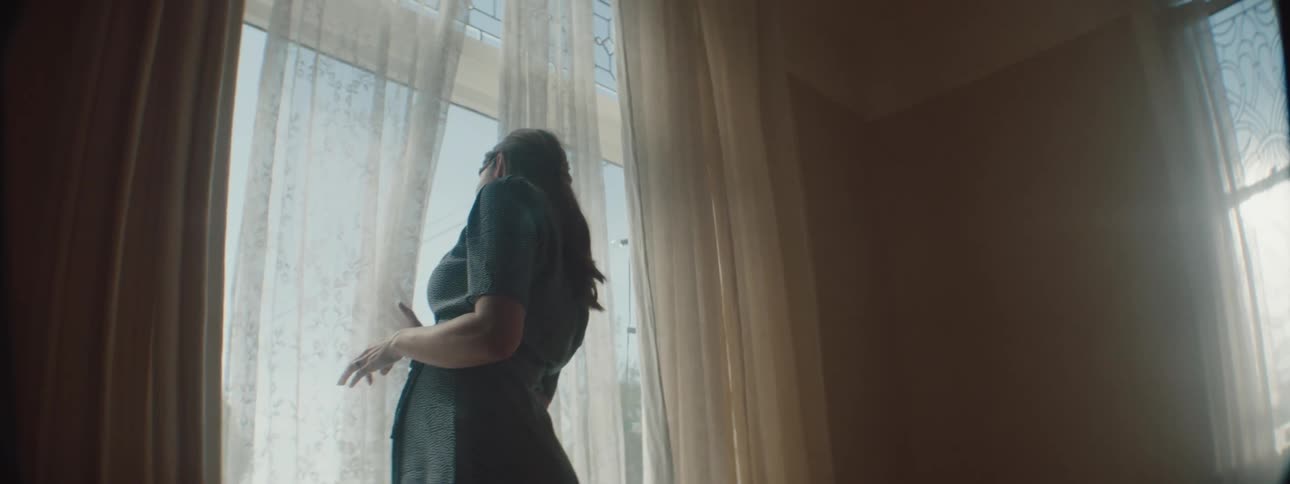 Woman with blue dress and brown hair touches the white window curtains