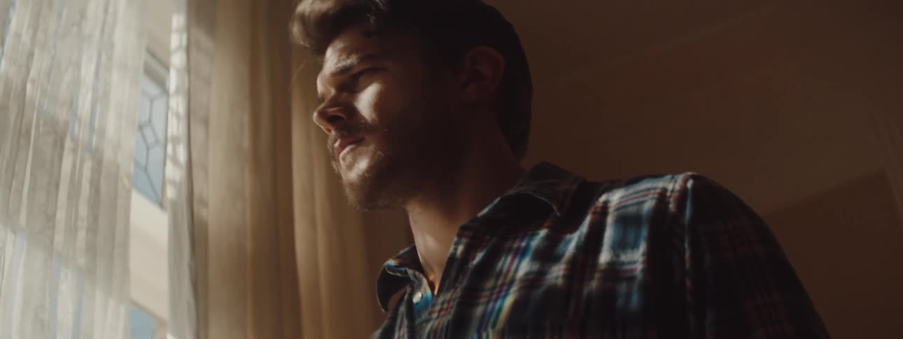 Bearded man in a plaid shirt stands indoors and looks out a window