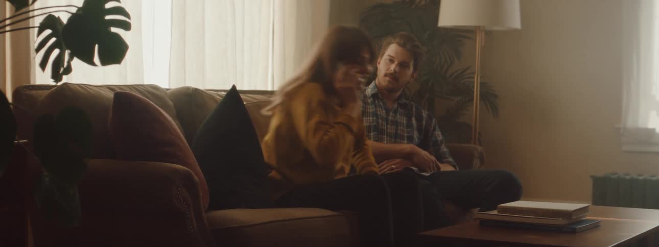 A man and woman sit together on the couch and then the woman picks up a cell phone from the table and starts talking