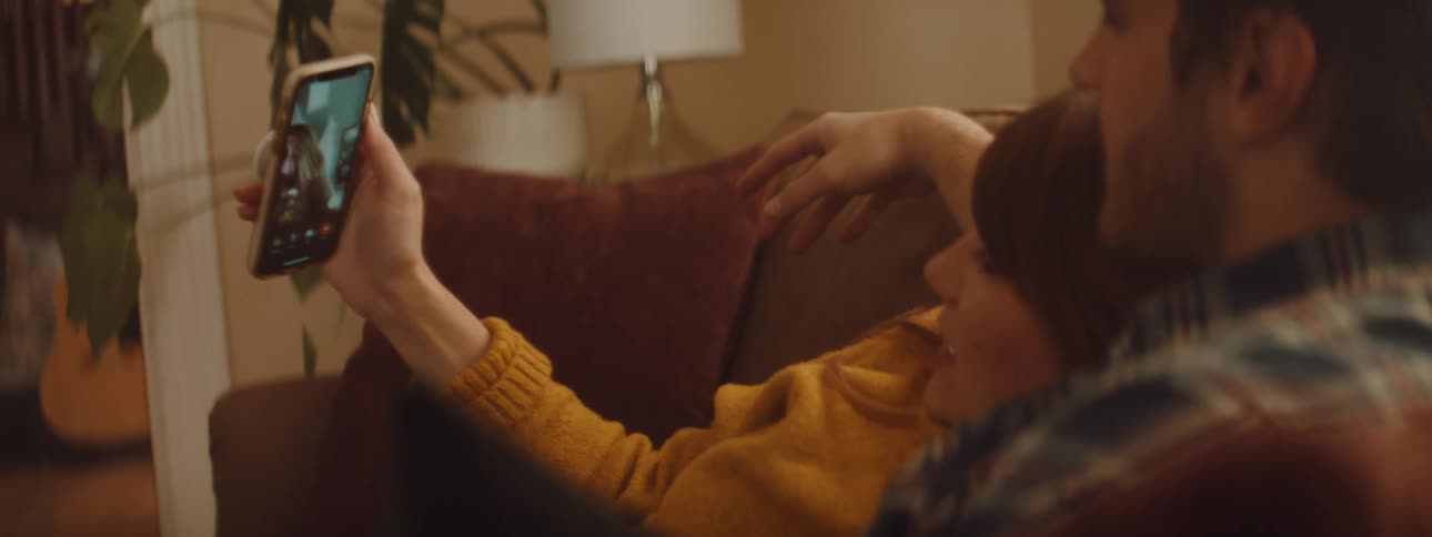 A man and woman cuddle on a couch and video caht with someone on a cell phone