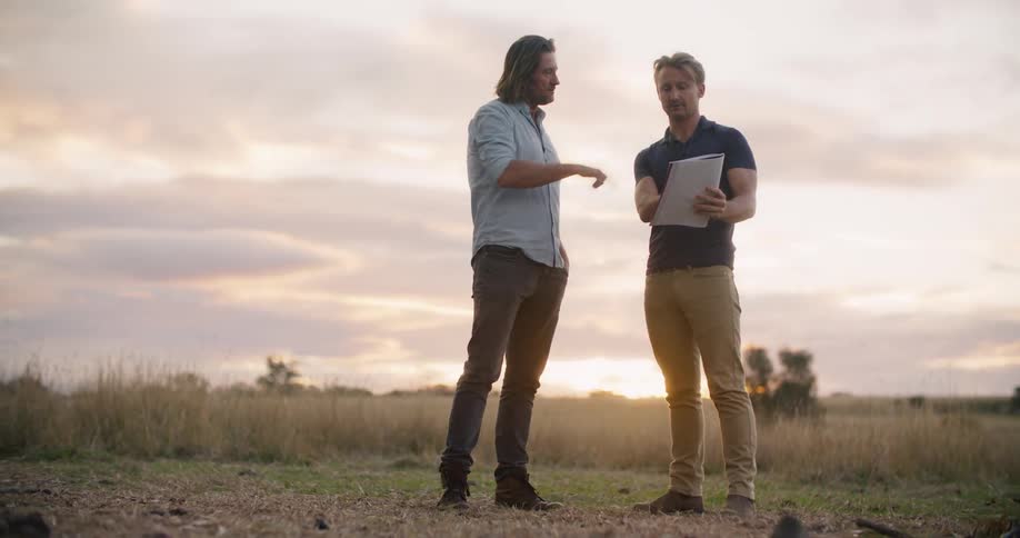 Two men stand in a field while one man holds a notebook
