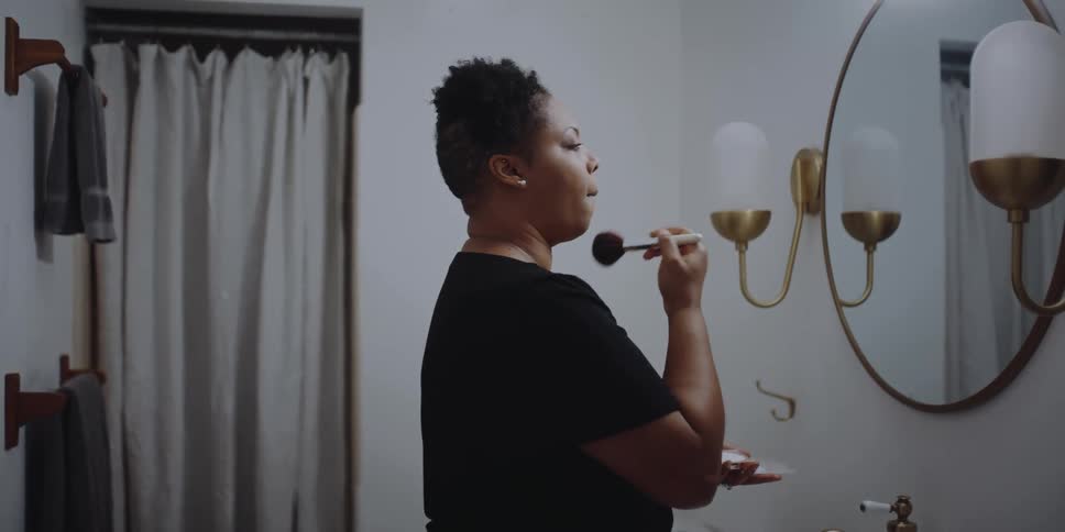 A woman in a room in front of a mirror putting on makeup with a makeup brush