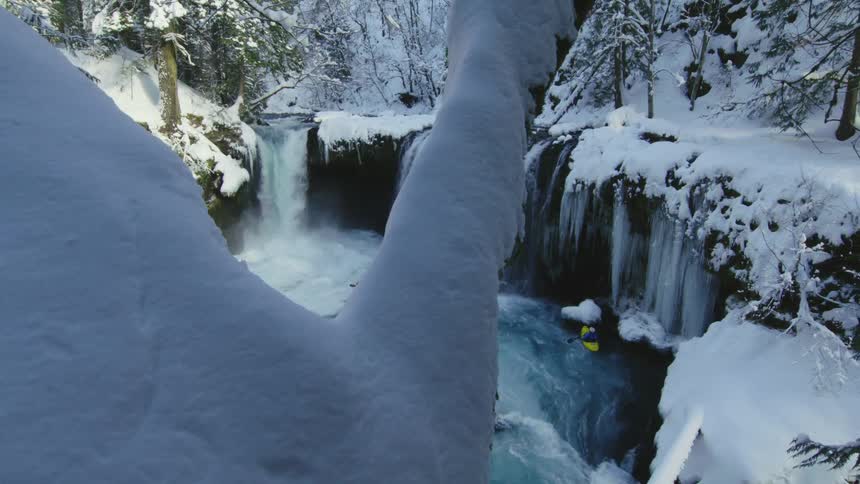 Slow pan to the right showing an incredible winter scene of icicles, pristine river water and three whitewater kayakers descending a perfect waterfall in heavy snow