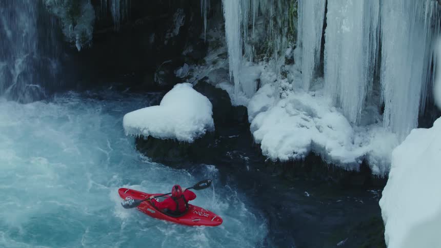 A man in a red whitewater kayak paddles next to huge frozen icicles on a teal pristine river on New Years Eve deep in the wilderness