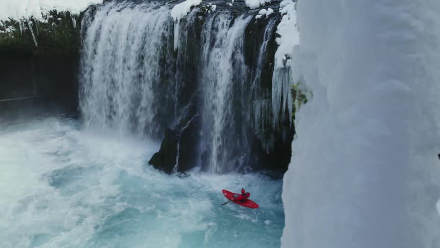A man in a red whitewater kayak paddles at the base of a huge waterfall and icicles in the dead of winter after a heavy snow and cold snap froze the winter wonderland