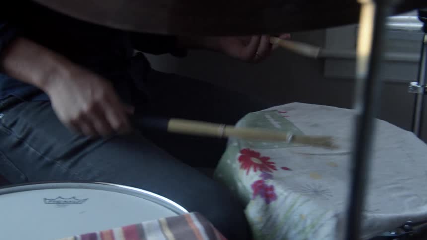 A man in a room sitting and playing drums with drumsticks with a wall and window in the background