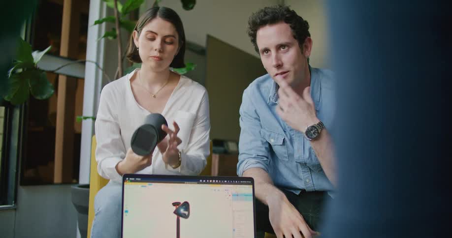 A man and a woman sitting in a living room with a small group of people surrounding a laptop passing a lamp to one another