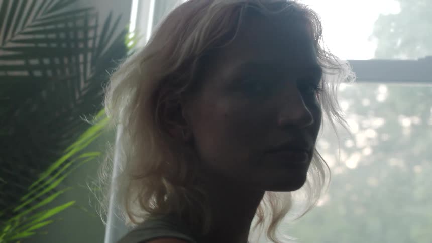 A woman in a room staring with plants a wall and a window in the background