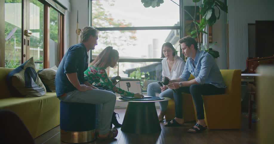 Two men and two women sitting on couches and chairs looking at a laptop with a wall and window in the background