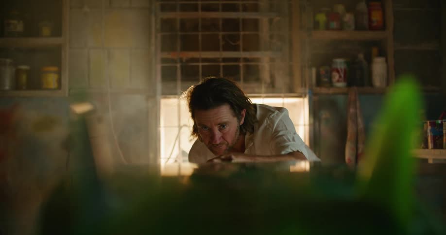 A man in a room looking at a surfboard with a wall and window in the background