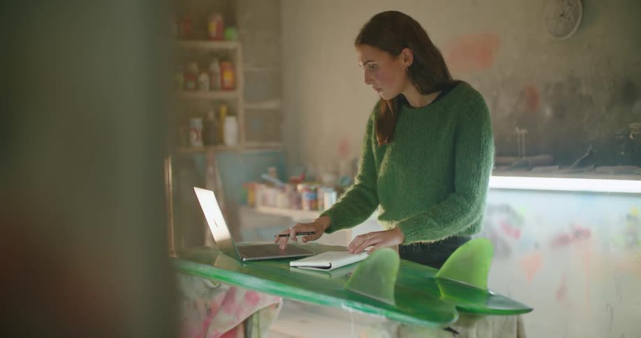 A woman uses a laptop resting atop a surfboard before stopping to write in a notebook