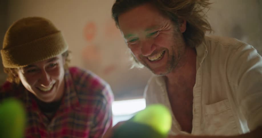 Older man smooths cloth over surfboard in workshop talking and smiling with younger man