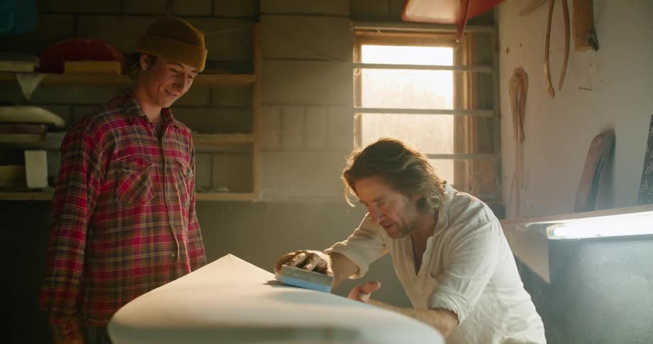 Two men in a room looking at a surfboard with a wall and window in the background