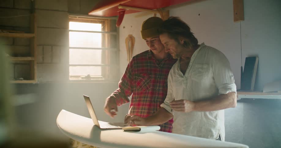 Father and son talking in front of a laptop placed on top of a surfboard inside a workshop