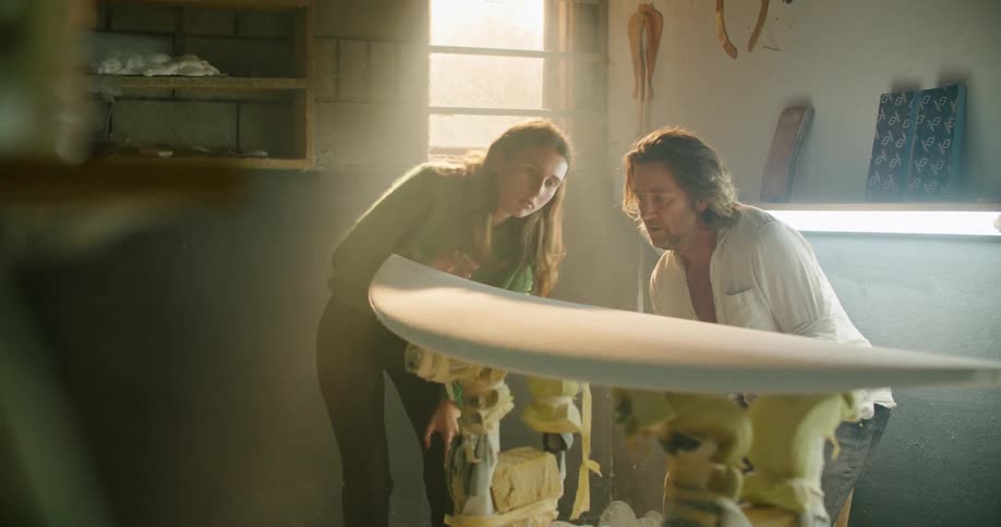A man brushes an unfinished surfboard in a workshop while showing it to a young woman who runs her hand along the surface