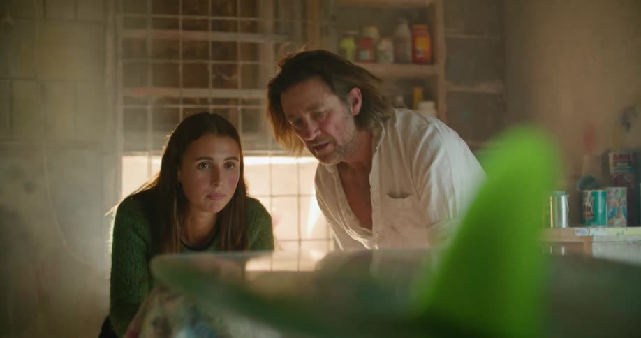 A man and a woman in a room looking at a surfboard with a wall and window in the background