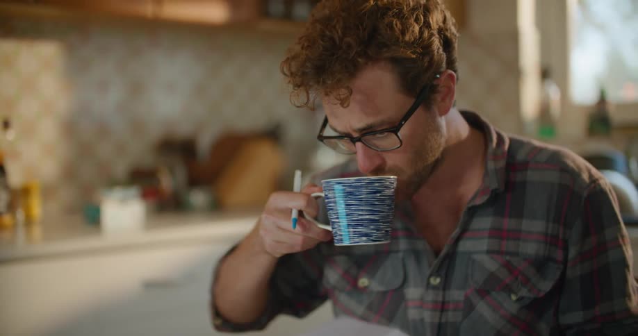 A man sitting at home in front of his computer picks up a cup of coffee and takes a drink