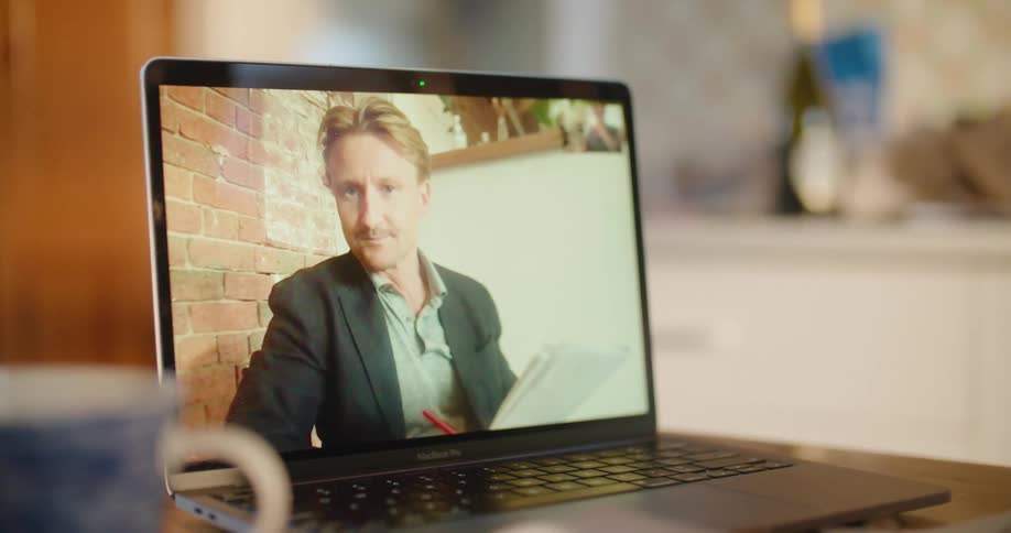Man on a laptop screen talks while holding paper and a pen