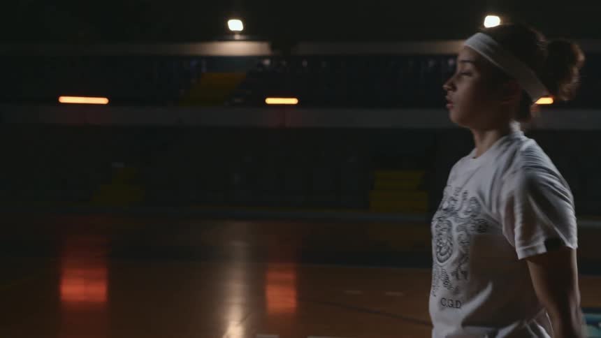 Girl wearing sweatband stands on basketball court with empty seats taking shots at hoop