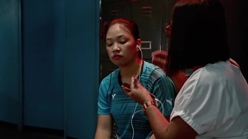 Two women sitting in the locker room listening to earbuds attached to a smart phone with lockers and locks in the background