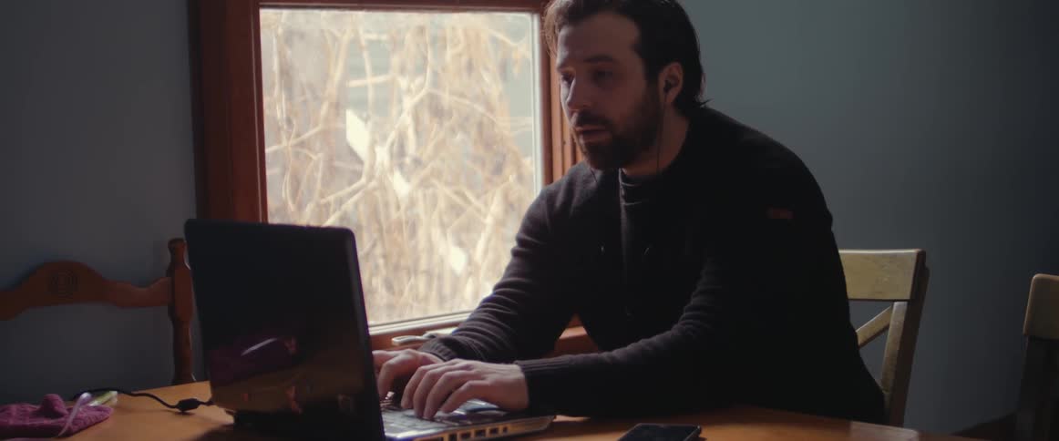A fearful man types on a laptop computer while sitting at a wooden table