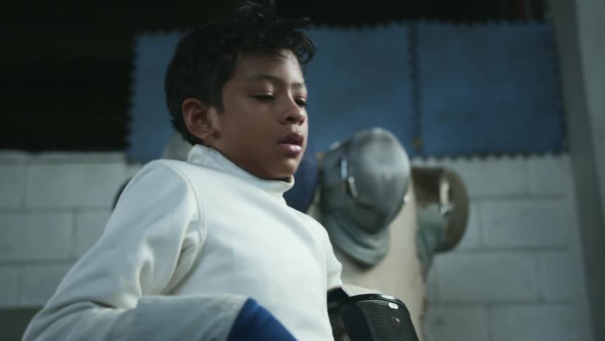 A boy removes his fencing helmet and takes a deep breath as his coach pats his chest